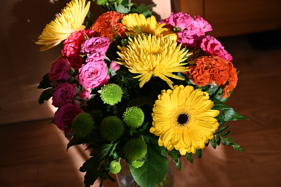 A vibrant bouquet of fresh flowers arranged in a glass vase, featuring large yellow gerbera daisies, bright yellow chrysanthemums, pink roses, orange carnations, and green button poms, accented with d