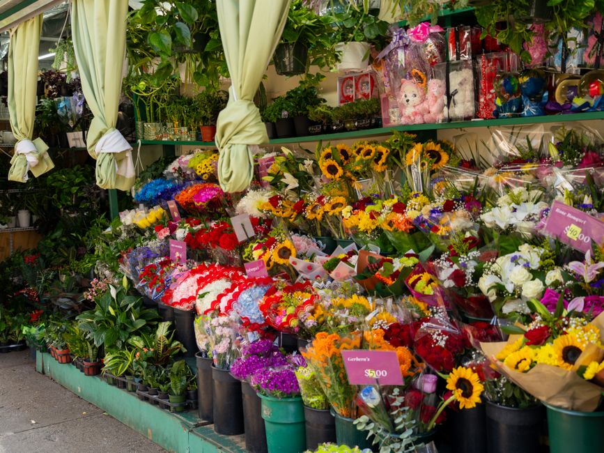 A vibrant display of fresh flower bouquets arranged on an outdoor stand, featuring a variety of colourful blooms including bright yellow sunflowers, red roses, purple statice, and white lilies, all wr