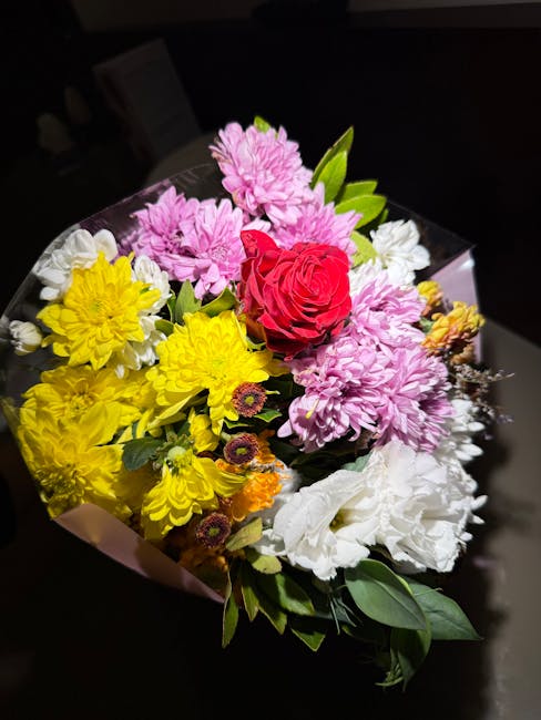 A vibrant floral arrangement featuring a central red rose surrounded by pink chrysanthemums, bright yellow daisies, white lisianthus, and small orange and purple filler flowers. The bouquet is wrapped