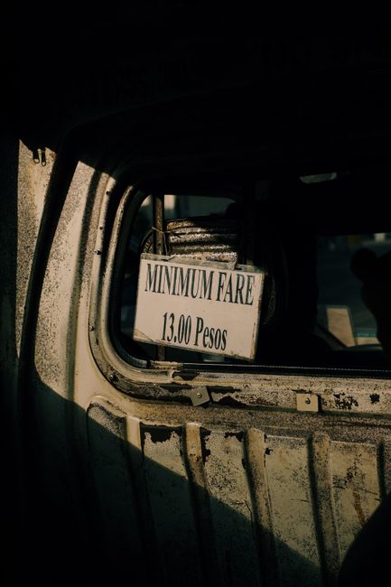 A close-up view of the inside of an old, weathered vehicle showing a rectangular sign in the rear window that reads 'MINIMUM FARE 13.00 Pesos.' The sign is white with black text, attached securely to 