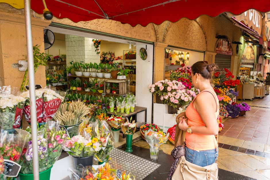 A woman in an orange tank top and jeans is standing outside a flower shop near Norbiton station, Kingston Vale, holding a small bouquet of flowers. The shop displays a variety of floral arrangements i