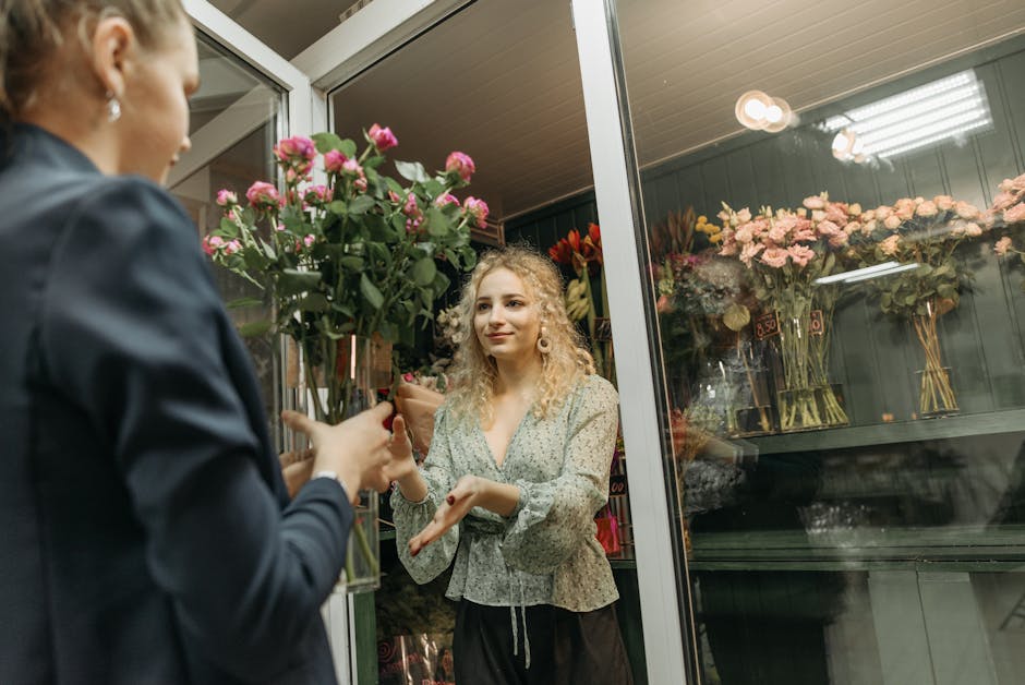 A female florist with curly blonde hair and a floral patterned blouse is receiving a bouquet of fresh pink roses with green foliage from a customer at the flower shop. The florist's hands are outstret