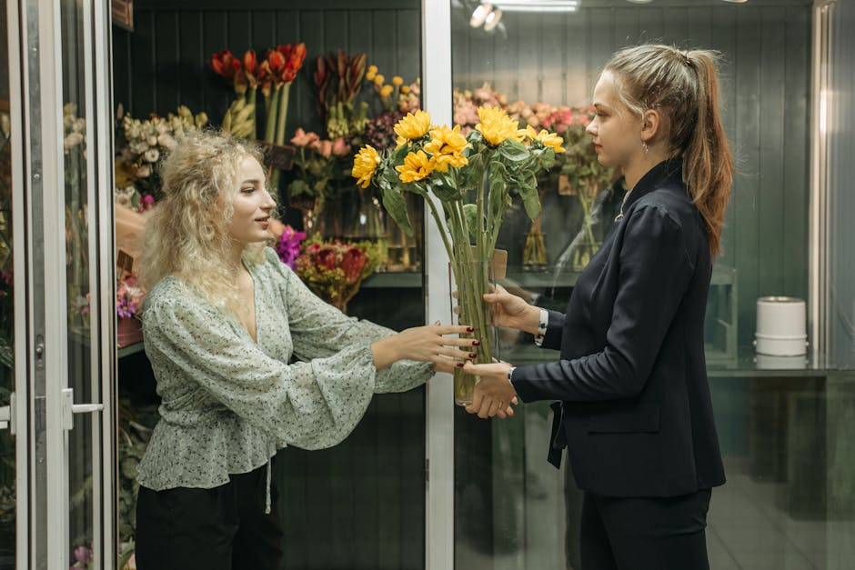 A woman with curly blonde hair and a light green floral blouse is receiving a fresh floral bouquet from a florist with long light brown hair tied back, dressed in a navy blazer. The bouquet features b
