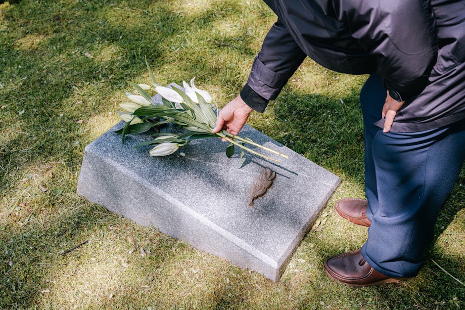 A person dressed in a dark jacket and navy trousers is placing a bouquet of fresh white lilies and greenery on a gray granite memorial stone at a cemetery. The bouquet is wrapped with a clear plastic 