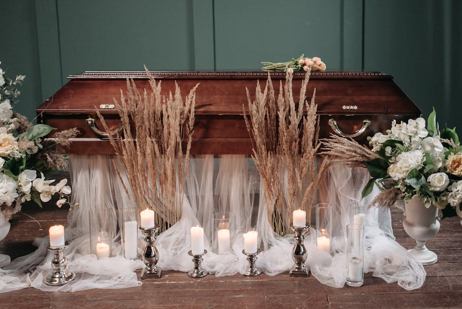 A wooden casket decorated with floral arrangements of white roses, peonies, and greenery on either side, set against a dark green wall. Surrounding the casket are tall, dried pampas grass stems and wh