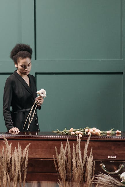 A young woman with dark skin and curly hair, dressed in a black blazer, stands in front of a dark green wall holding a single white flower with pink accents. In front of her, on a wooden table, lies a