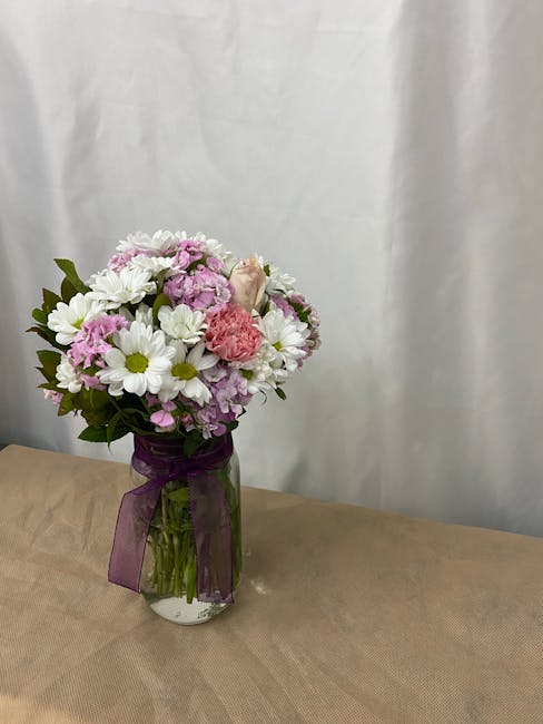 A floral bouquet arranged in a clear glass vase, featuring a mix of fresh flowers including white daisies with yellow centers, pink carnations, and pastel purple stocks, complemented by green foliage.