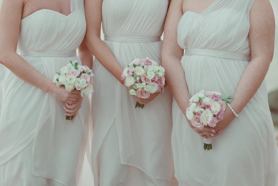 Three women dressed in elegant white wedding gowns holding small, round bouquets composed of white and soft pink roses, pastel pink ranunculus, and greenery accents. The bouquets are wrapped in light