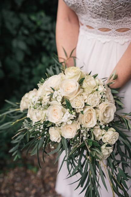 A close-up of a bridal bouquet featuring a lush arrangement of cream and ivory roses, complemented by small white filler flowers and long, slender green leaves, held by a bride dressed in a white lace