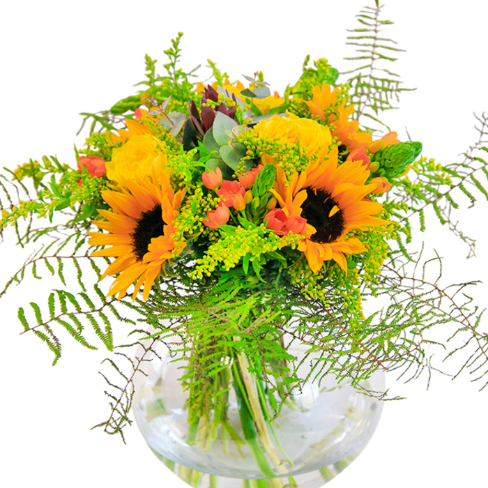 From the first glance, this bouquet radiates relentless optimism and summer abundance, echoing the landscape of Kingston Vale with its rolling greens and sunlit gardens. The composition is led by resplendent sunflowers that unfurl in bold, golden rays, their earthy brown centres evoking fields glowing under the wide Kingston Vale sky. Alongside, delicate yellow roses gently spiral open, their intricate petals layered in soft whorls-a reminder of quiet afternoons in the garden or leisurely walks along Kingston Hill. Glossy clusters of red hypericum berries are scattered throughout, adding an unexpected burst of energy amidst subtle greenery. Lush, feathery ferns and sprigs of wispy foliage spill outward in relaxed curls, softening the bouquet's contours and imbuing it with a sense of effortless movement. Hints of blue-green eucalyptus thread their way through the arrangement, providing a cool contrast to the warmth and a trace of fresh, herbal scent. The whole composition is cradled in a spherical, crystal-clear glass vase, where the submerged stems are neatly displayed in fresh water-a detail that speaks to the care and artistry of hand-delivered bouquets from Kingston Vale's expert florists. Bathed in natural light, the bouquet's textures-plush petals, sleek berry skins, and crisp foliage-invite closer inspection. Ideal for joyful occasions or as a statement of warmth in a local home, this arrangement captures the spirit of celebration and hospitality that defines Kingston Vale.